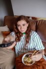 Von oben junge rothaarige Frau, die Ramen isst und Kanäle im Fernsehen wechselt, während sie zu Hause auf dem Sofa sitzt — Stockfoto