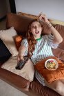 D'en haut jeune rousse femme manger ramen et changer de chaînes à la télévision tout en étant assis sur le canapé pendant le déjeuner à la maison — Photo de stock