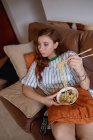 From above young redhead woman eating ramen and changing channels on TV while sitting on sofa during lunch at home — Stock Photo