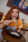 From above young redhead woman in casual clothes using chopsticks while sitting at table and eating sushi at home — Stock Photo