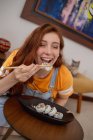 D'en haut jeune rousse femme en vêtements décontractés en utilisant des baguettes tout en étant assis à la table et en mangeant des sushis à la maison — Photo de stock