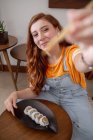 Зверху молода руда жінка в повсякденному одязі дивиться на камеру, використовуючи палички, сидячи за столом і їдячи суші вдома — стокове фото
