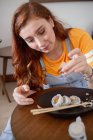 Сверху молодая рыжая женщина в повседневной одежде палочками для еды, сидя за столом и поедая суши дома. — стоковое фото