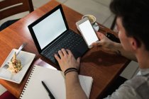 Von oben sitzt ein Mann am Tisch und surft Laptop und Smartphone mit leeren Bildschirmen, während er im Café an einem Remote-Projekt arbeitet — Stockfoto