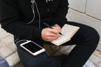 Desde arriba chico anónimo en ropa casual escuchando música en el teléfono inteligente y tomando notas en el bloc de notas mientras está sentado en la calle de la ciudad - foto de stock