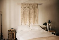Винтажное украшение макраме висит на стене над удобной кроватью в уютной спальне дома — стоковое фото