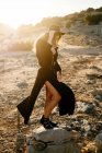 Senhora da moda de comprimento total em roupa de país preto ajustando chapéu e de pé sobre pedra durante o pôr do sol na natureza — Fotografia de Stock