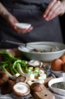 Bando de cebolinhas frescas e cogumelos colocados na tábua de corte perto de ovos e sementes de papoula contra a dona de casa da colheita misturando ingredientes na tigela — Fotografia de Stock