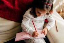 Nettes kleines Mädchen sitzt auf der Couch und zeichnet im Notizbuch — Stockfoto