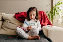 Cute little girl sitting on the couch and drawing in the notebook — Stock Photo
