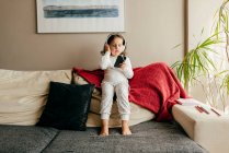 Cute little girl sitting on the couch listening music with headphones — Stock Photo