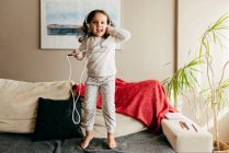 Cute little girl jumping on the couch and listening music with headphones — Stock Photo
