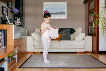 Menina bonito em leotard rosa claro e meia calça vestindo saia enquanto está perto de sapatos de dança na acolhedora sala de estar em casa — Fotografia de Stock