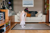 Симпатична маленька дівчинка в світло-рожевому леопарді і колготках одягається на спідницю, стоячи біля танцювального взуття в затишній вітальні вдома — стокове фото