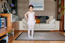 Menina bonito em leotard rosa claro e meia calça vestindo saia enquanto está perto de sapatos de dança na acolhedora sala de estar em casa — Fotografia de Stock