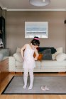Cute little girl in light pink leotard and tights putting on skirt while standing near dance shoes in cozy living room at home — Stock Photo