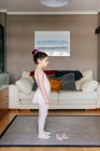 Menina bonito em leotard rosa claro e meia-calça se preparando enquanto está perto de sapatos de dança na acolhedora sala de estar em casa — Fotografia de Stock