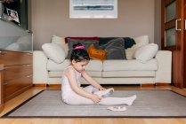 Vista lateral da menina bonito em collants e collants sentado no chão perto do sofá e vestindo sapatos de dança antes do ensaio de balé em casa — Fotografia de Stock
