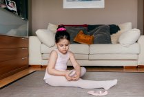 Side view of cute girl in leotard and tights sitting on floor near sofa and putting on dance shoes before ballet rehearsal at home — Stock Photo