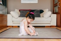 Vista lateral de la linda chica en maillot y medias sentadas en el suelo cerca del sofá y poniéndose zapatos de baile antes del ensayo de ballet en casa - foto de stock