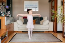 Linda niña con los brazos extendidos mirando hacia otro lado y bailando cerca del sofá durante el ensayo de ballet en casa - foto de stock