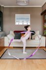 Вид сбоку на милую маленькую девочку в трико и трико, крутящуюся ленту и танцующую во время художественной гимнастики в уютной гостиной дома — стоковое фото