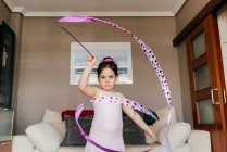 Focused cute little brunette girl in leotard looking at camera while spinning ribbon during rhythmic gymnastic practice training in cozy living room at home — Stock Photo