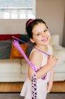 Vue latérale de mignonne petite fille brune heureuse avec ruban souriant regardant la caméra pendant l'entraînement gymnastique rythmique à la maison — Photo de stock
