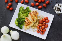 Vue du dessus de délicieux poulet frit et brocoli placés sur une assiette près des oignons et des tomates cerises sur une table grise près du poivre — Photo de stock