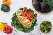 Dall'alto pollo arrosto con broccoli, pepe rosso e avocado dimezzato ono un letto di foglie verdi in un tavolo di marmo — Foto stock