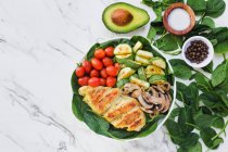 From above cherry tomatoes placed inside bowl of roasted chicken and zucchini with mushrooms on spinach leaves near half avocado and spices — Stock Photo