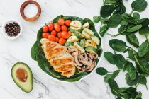 From above cherry tomatoes placed inside bowl of roasted chicken and zucchini with mushrooms on spinach leaves near half avocado and spices — Stock Photo