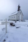 Bajo ángulo de la pequeña iglesia del pueblo situada cerca del cementerio local con la cruz blanca entre el campo cubierto de nieve contra el cielo gris nublado en el día de invierno en Islandia - foto de stock