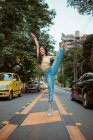 Woman in casual clothes doing splits with raised arm and smiling while dancing on asphalt road amidst cars on busy street of modern city — Stock Photo
