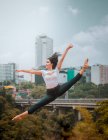 Босоногая женщина в повседневной одежде прыгает и делает сплит, танцуя против современного города и облачного неба — стоковое фото