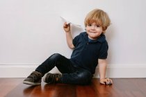 Netter blonder Junge in lässigem Outfit sitzt auf Holzboden und spielt mit Papierflieger und lächelt fröhlich in die Kamera auf weißem Wandhintergrund — Stockfoto