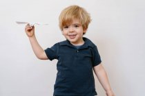Bonito menino loiro em roupa casual brincando com o avião de papel sorrindo brilhantemente para a câmera no fundo da parede branca — Fotografia de Stock