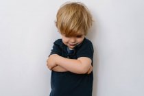 Annoyed blond little boy in casual clothes looking down with dissatisfaction while standing on white wall with crossed arms — Stock Photo