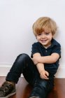 Adorabile ragazzo prescolare in maglietta casual sorridente guardando altrove seduto in un pavimento di legno e appoggiato su uno sfondo bianco della parete — Foto stock