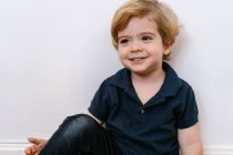Adorable garçon d'âge préscolaire en tee-shirt décontracté souriant regardant loin assis et penché dans un fond mural blanc — Photo de stock