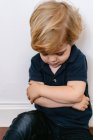 Раздраженный блондин маленький мальчик в повседневной одежде сидит на полу со скрещенными руками — стоковое фото
