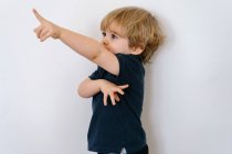Adorable garçon d'âge préscolaire en tee-shirt décontracté regardant loin tout en levant le bras en jouant à des jeux de doigts penché dans un fond de mur blanc — Photo de stock