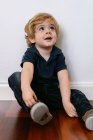 Menino pré-escolar adorável em camiseta casual sorrindo olhando para longe sentado em um chão de madeira e inclinado em um fundo de parede branca — Fotografia de Stock