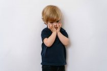 Infastidito bambino biondo in abiti casual guardando lontano con insoddisfazione mentre in piedi su una parete bianca con le mani sul viso — Foto stock