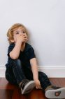 Menino pré-escolar adorável em camiseta casual olhando para longe sentado em um chão de madeira e inclinado com a mão cobrindo a boca no fundo da parede branca — Fotografia de Stock