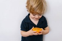 Engraçado menino loiro em roupas casuais comendo metade de laranja olhando para longe com o sorriso enquanto em pé contra o fundo branco — Fotografia de Stock