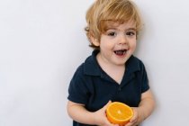 Смешной блондин маленький мальчик в повседневной одежде ест половину апельсина глядя в сторону с улыбкой, стоя на белом фоне — стоковое фото