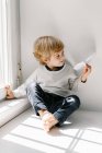 Glückliches blondes kleines Kind in lässiger Kleidung, das an sonnigen Tagen barfuß auf der Fensterbank sitzt und mit Papierflugzeug spielt — Stockfoto