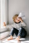 Criança loira feliz em roupas casuais brincando com avião de papel enquanto sentado descalço no peitoril da janela no dia ensolarado — Fotografia de Stock