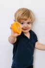 Petit garçon blond drôle en vêtements décontractés s'étendant offrant la moitié de l'orange vers la caméra et sortant la langue avec le sourire tout en se tenant sur fond blanc — Photo de stock